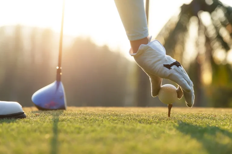 En golfspiller iført hanske plasserer en golfball på en tee på Golf Innlandet, med en golfkølle og den solfylte banen i bakgrunnen.