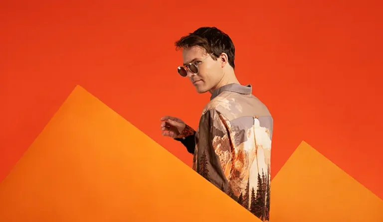 En mann i solbriller står foran en oransje bakgrunn på Klubbkveld med Standup Innlandet.