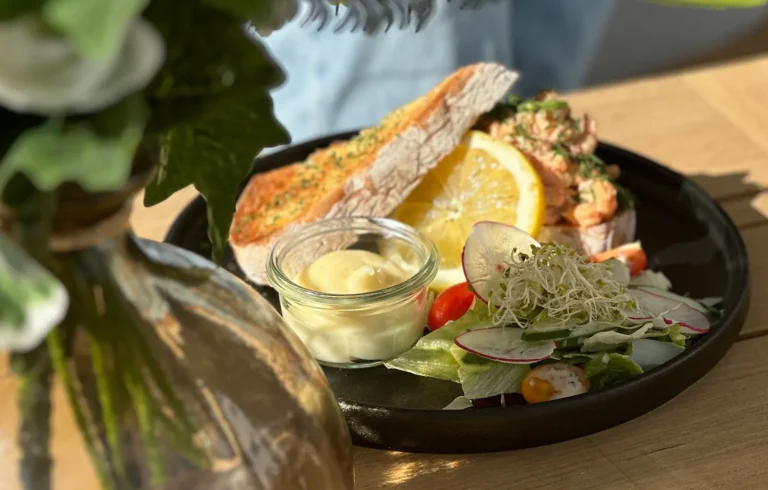 En tallerken med ristet smørbrød, sitronskive, en liten glassbolle med saus, og en sidesalat med cherrytomater og spirer på 17 mai på Central Restaurant & Bar. En blomstervase er delvis synlig i forgrunnen.