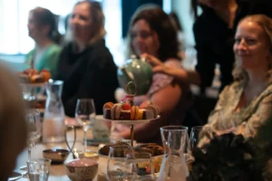 En gruppe mennesker sitter rundt et bord på Central Restaurant og Bar og nyter et brett med diverse bakverk og drikkevarer, mens noen skjenker te fra en tekanne i bakgrunnen.