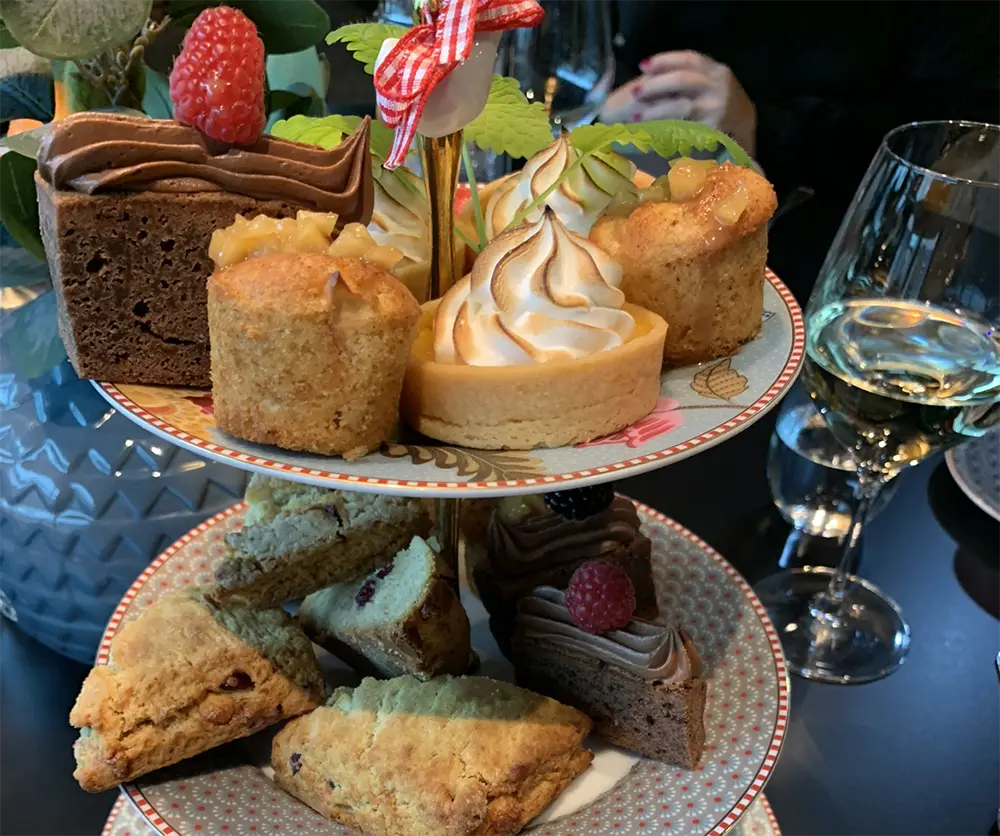 Et utvalg desserter, inkludert sjokoladekake, sitronterte og scones arrangert på et to-etasjes serveringsbrett, med vinglass og en potteplante i bakgrunnen – perfekt for en jul Afternoon Tea lørdag 7. desember.