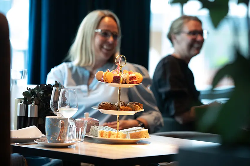 To kvinner sitter ved et bord på Hotel Central og nyter en ettermiddagste. Bordet har et brett med bakverk, scones og smørbrød, samt tekopp, fat, vinglass til smaking av sommerens bobler og noen småretter.