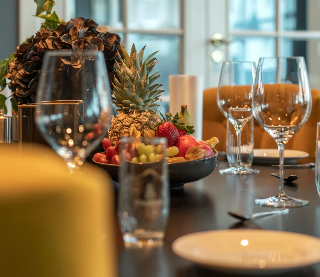 Elegant spisebord til Åpningsmiddag onsdag 13. november, med tomme vinglass, en rekke frukt inkludert en ananas og jordbær i en bolle, supplert med gule polstrede stoler.