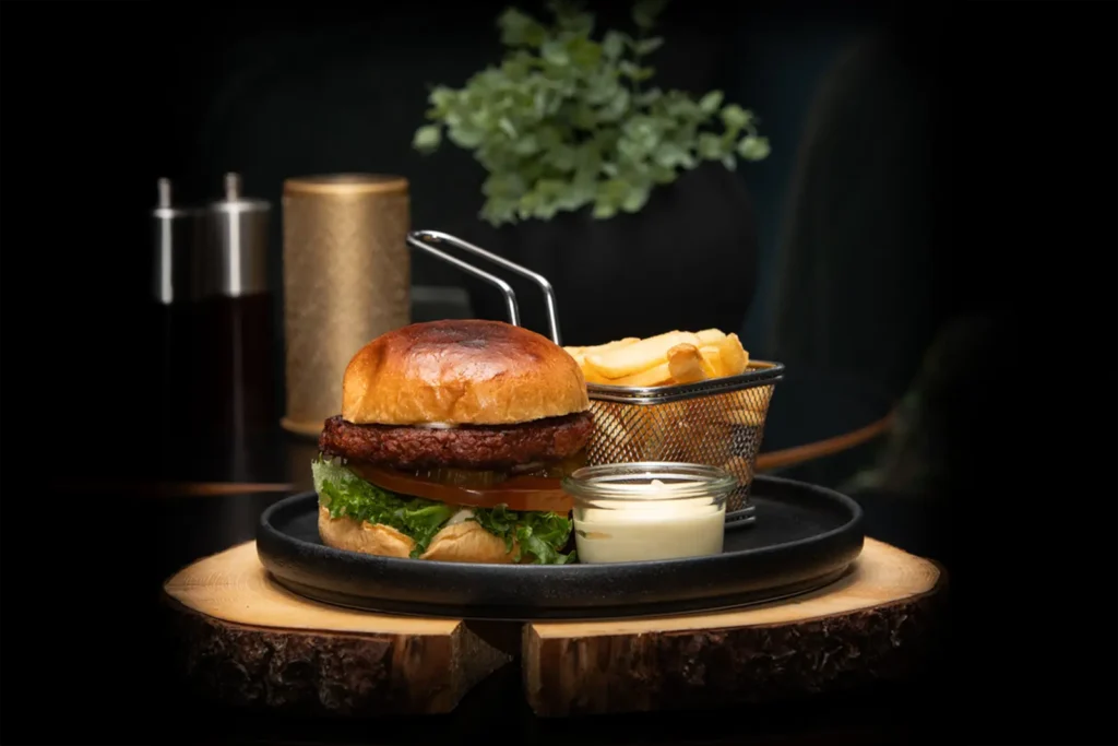 En gourmetburger med salat og tomat, servert med pommes frites i en liten kurv og en sausbeholder, satt på en svart tallerken med underlegg av tre og en plante i bakgrunnen. Bli med oss på denne herlige opplevelsen på Serata de Festa på Central Restaurant 17. oktober.