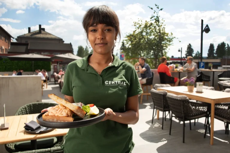 En servitør i grønn poloskjorte med «Central»-logo bærer en tallerken med mat på den livlige uteplassen den 17. mai på Central Restaurant & Bar, mens gjestene nyter den festlige atmosfæren ved bordene i nærheten.