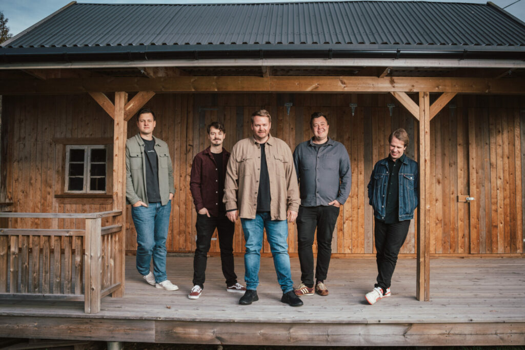 Fem menn står avslappet på verandaen til en rustikk hytte, alle vendt mot kamera iført jakker og jeans - og fanger en avslappet stemning som minner om en kveld på wig wam på Central Scene.