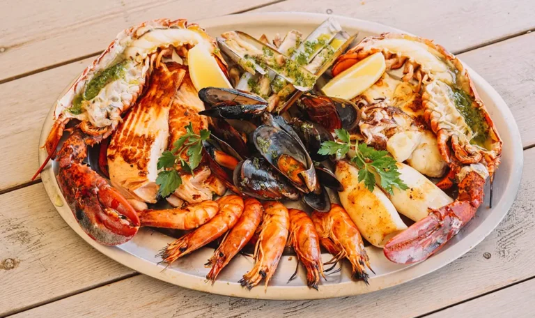 Et fat med assorterte skalldyr, inkludert hummer, reker, blåskjell, fiskefileter og knivmuslinger, garnert med sitronbåter og persille på et trebord.
