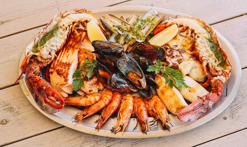 Et fat med assorterte skalldyr, inkludert hummer, reker, blåskjell, fiskefileter og knivmuslinger, garnert med sitronbåter og persille på et trebord.