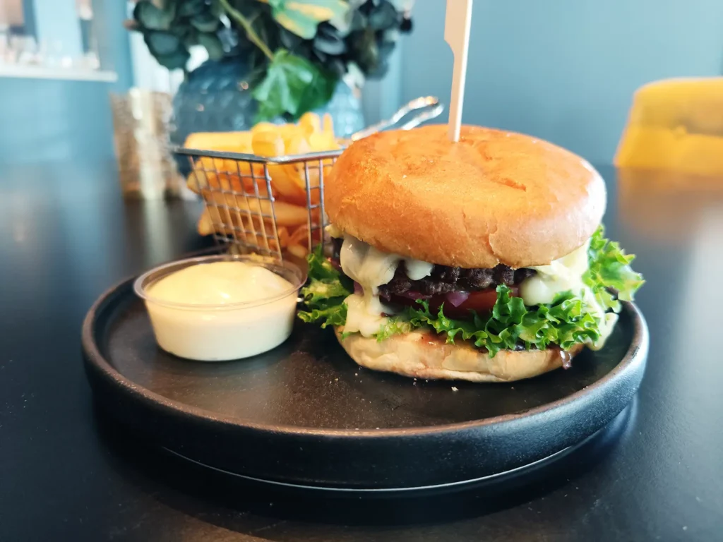 På Restaurant Elverum serveres en klassisk cheeseburger med salat og tomat på et brød, sammen med sprø pommes frites i en metallkurv og et lite beger med dippesaus på en svart tallerken.