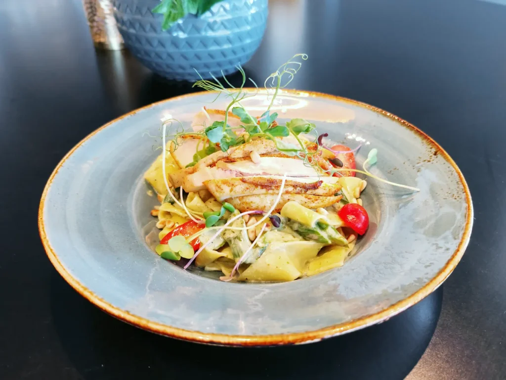 En tallerken pasta med grillet kylling, cherrytomater og mikrogrønt, servert i en grå keramikkskål på et mørkt bord på restaurant Elverum.