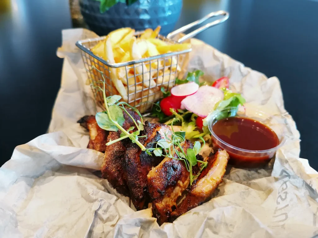 En porsjon grillet ribbe med mikrogrønt, pommes frites i metallkurv, salat med reddiker og en kopp barbecuesaus på pergamentpapir på Restaurant Elverum.