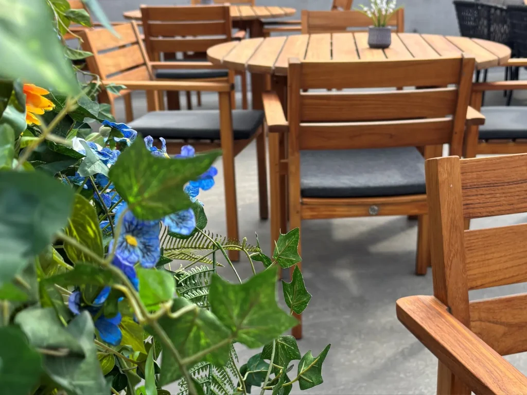 Flere trebord og stoler med grå puter er plassert på en uteplass på Restaurant Elverum. I forgrunnen skimtes lilla blomster og grønne blader.
