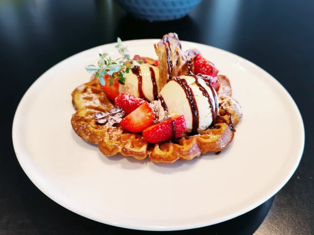 På Restaurant Elverum venter en vaffel toppet med to kuler vaniljeis, jordbær i skiver, sjokoladesaus, melis og en urtekvist på en hvit tallerken.