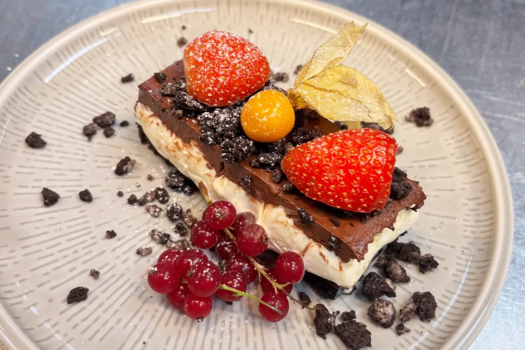 På restaurant Elverum kan du nyte en rektangulær dessert toppet med sjokolade, jordbær, et gyllent bær, rips, smuldrede småkaker og melis på en beige tallerken.