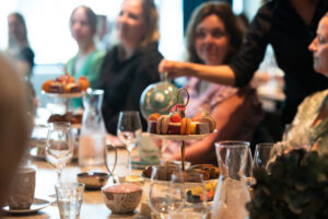 Folk sitter rundt et bord dekket med desserter og drikke på restaurant Elverum mens en person skjenker te fra en tekanne i glass.