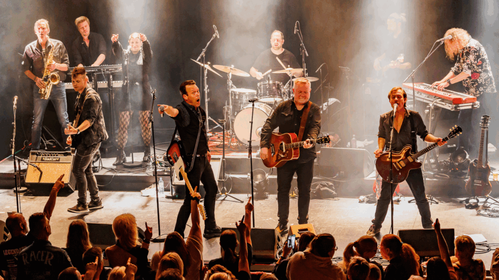 Et band med flere musikere og sangere opptrer på scenen med gitarer, saksofon, trommer og keyboard på wig wam på Central Scene, mens et publikum ser på fra forgrunnen.