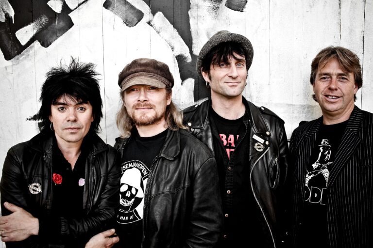 Fire menn i mørke klær og jakker står foran en graffitikledd vegg og poserer for et gruppebilde etter arrangementet Wig Wam på Central Scene.