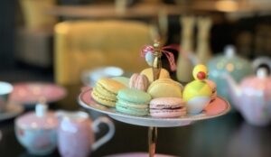 Et brett med ulike pastellfargede makroner og pyntegjenstander er perfekt for en koselig Mandagsklubben Quizliga-samling, med tekopper og tekanne uskarpt i bakgrunnen.