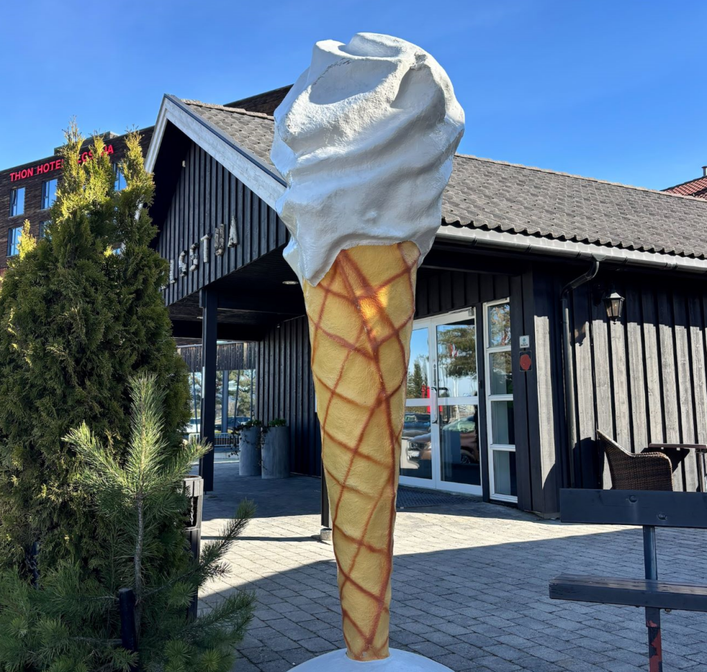 En stor iskremstatue står utenfor Elgstua Kafés mørke trebygning på en solrik dag, med trær og et hotellskilt i bakgrunnen.